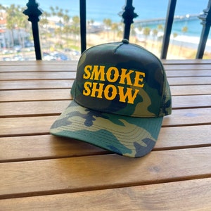 May include: A camouflage trucker hat with a yellow embroidered logo that says "SMOKE SHOW".