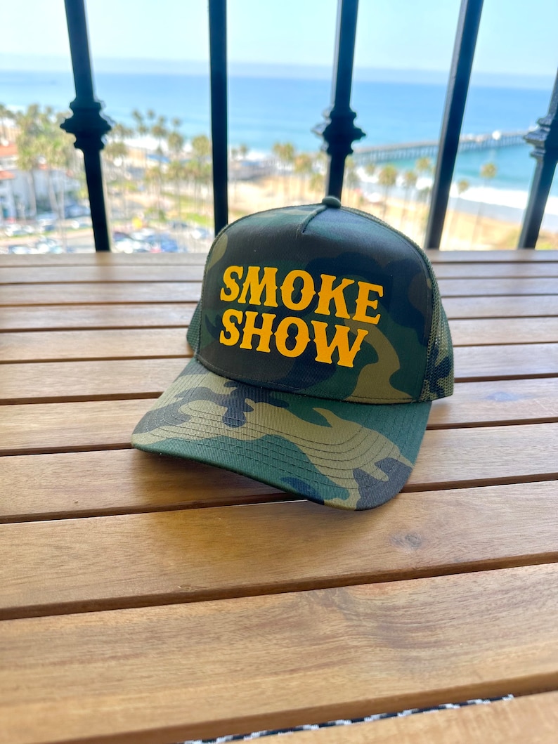 May include: A camouflage trucker hat with a yellow embroidered logo that says "SMOKE SHOW".