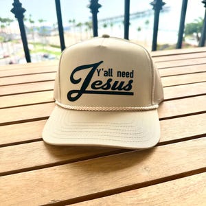 May include: Tan trucker hat with a mesh back and a curved brim. The front of the hat features the text "Y'all need Jesus" in a black script font. A rope detail accents the brim.