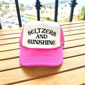 May include: A pink and white trucker hat with black embroidered text that reads "Seltzers and Sunshine".