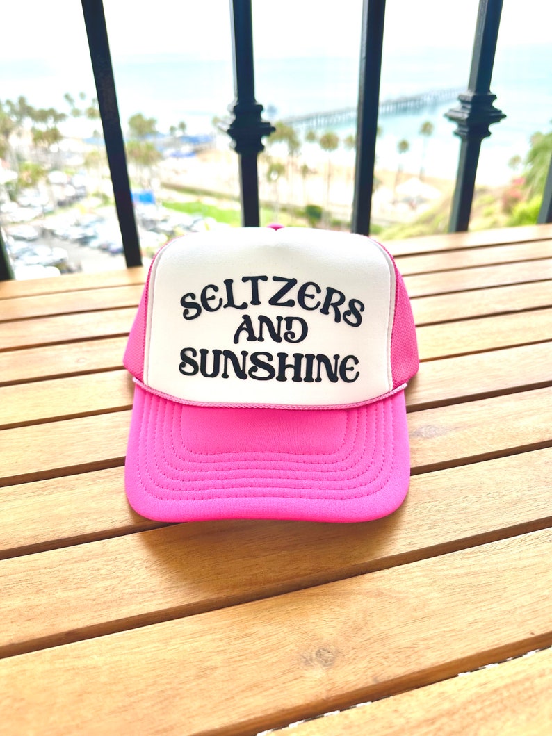 May include: A pink and white trucker hat with black embroidered text that reads "Seltzers and Sunshine".