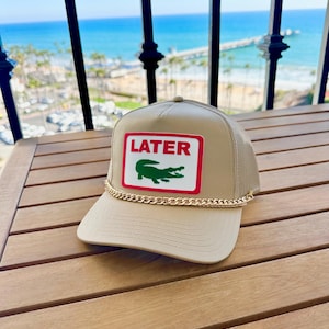 Pode incluir: Boné trucker bege com parte traseira em rede e aba curva. O patch frontal apresenta a palavra "LATER" em vermelho, com um logotipo de crocodilo verde. Um detalhe de corrente dourada adorna a aba.