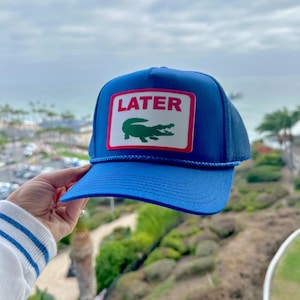 May include: Blue trucker hat with a curved brim and mesh back. The front features a rectangular patch with the word "LATER" in pink and a green crocodile logo. A blue rope detail accents the brim.