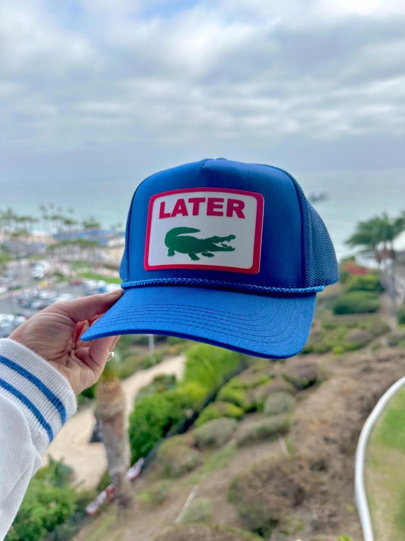 May include: Blue trucker hat with a curved brim and mesh back. The front features a rectangular patch with the word "LATER" in pink and a green crocodile logo. A blue rope detail accents the brim.