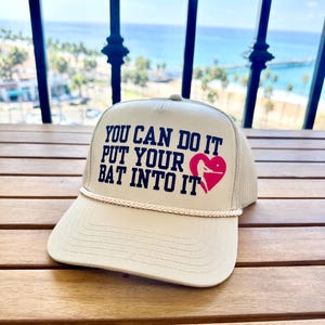 Puede incluir: Una gorra de béisbol beige con un diseño bordado en blanco y negro que dice "You can do it put your bat into it" con un corazón rojo y una silueta de jugador de béisbol dentro del corazón.