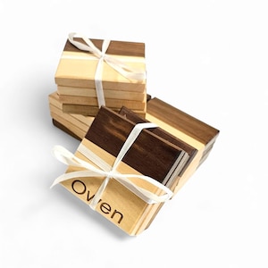 Puede incluir: Conjunto de cuatro posavasos de madera con un acabado de madera natural y un acento de madera oscura. Los posavasos están apilados y atados con una cinta blanca. El posavasos superior tiene el nombre "Owen" grabado.