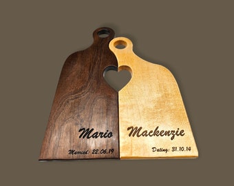 May include: Two wooden cutting boards, one dark brown and one light brown, interlock to form a heart shape. The dark board is engraved with "Mario" and "Married: 22.06.19". The light board reads "Mackenzie" and "Dating: 31.10.14".