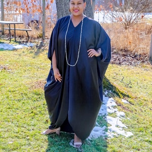 May include: A woman wearing a black, long-sleeved, flowing dress with a pearl necklace and a bracelet. She is standing in a grassy area with a tree behind her.