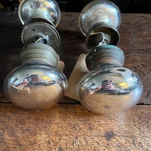 Könnte beinhalten: Zwei antike, silberfarbene Türknöpfe mit einem reflektierenden, kugelförmigen Design. Die Knöpfe weisen Gebrauchsspuren auf. Sie sind auf einer Holzoberfläche montiert, was auf eine rustikale Ästhetik hindeutet.