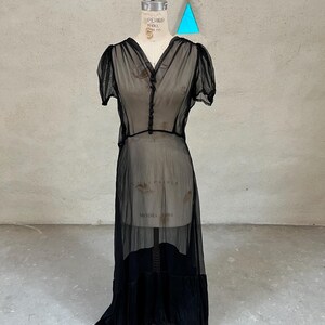Può includere: Un abito vintage nero trasparente con maniche corte e scollo a V. L'abito è esposto su un manichino, che mostra il suo design fluido e a strati. L'abito ha un davanti abbottonato e un orlo lungo e asimmetrico.