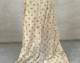 May include: A long, cream-coloured skirt featuring a repeating pattern of small black triangles and dots. The skirt has a brown waistband and a slight flare at the hem, creating a flowing silhouette. The fabric appears to be a lightweight material.