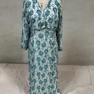 Op de afbeelding: Een vintage lichtblauwe jurk met een bloemenpatroon in teal en zilver. De jurk heeft lange mouwen en een V-hals. De jurk wordt op een mannequin getoond.