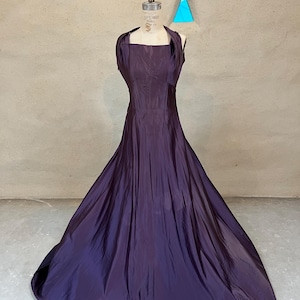 May include: A long, dark purple dress with a halter neck is shown on a dress form. The dress features a fitted bodice and a full, flowing skirt. The fabric has a subtle sheen, and the dress appears to be made of a lightweight material.