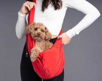 May include: A person carrying a small brown dog in a red sling carrier. The sling is worn over the person's shoulder and has a zipper closure.