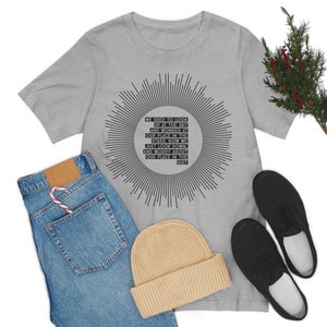 May include: A light gray t-shirt with a black sunburst design and text that reads "WE USED TO LOOK UP AT THE SKY AND WONDER AT OUR PLACE IN THE STARS. NOW WE JUST LOOK DOWN, AND WORRY ABOUT OUR PLACE IN THE DIRT." The shirt is paired with blue jeans, a tan beanie, and black shoes.