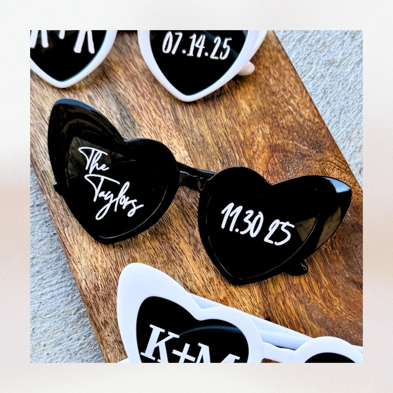 May include: Black heart-shaped sunglasses with white text that reads "The Taylors" on one lens and "11.30.25" on the other lens.  The sunglasses are on a wooden surface.