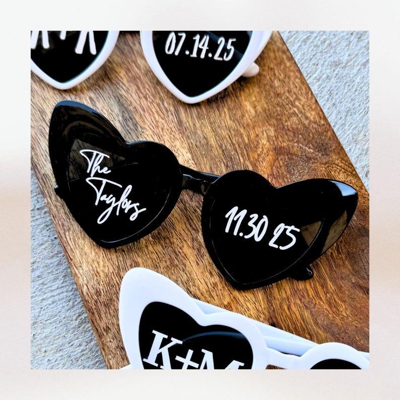 May include: Black heart-shaped sunglasses with white text that reads "The Taylors" on one lens and "11.30.25" on the other lens.  The sunglasses are on a wooden surface.