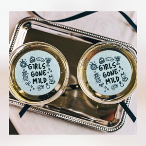 K&ouml;nnte beinhalten: Zwei Champagnerfl&ouml;ten mit goldenen R&auml;ndern, jede mit einem wei&szlig;en Papieruntersetzer, auf dem in schwarzer Schrift "Girls Gone Mild" steht. Die Untersetzer sind mit kleinen schwarzen Illustrationen von partybezogenen Gegenst&auml;nden verziert.