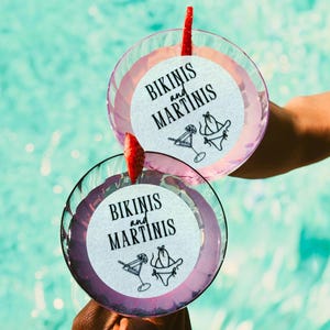 K&ouml;nnte beinhalten: Zwei rosa Cocktails mit wei&szlig;en Papieruntersetzern, auf denen in schwarzer Schrift "Bikinis and Martinis" steht. Die Untersetzer zeigen au&szlig;erdem eine Schwarzwei&szlig;illustration eines Bikinis und eines Martiniglases.