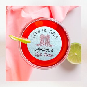 K&ouml;nnte beinhalten: Ein runder Aufkleber in Blau und Wei&szlig; mit dem Text "Let's Go Girls" und einem Paar Cowboystiefeln. Der Aufkleber befindet sich auf einem roten Getr&auml;nk in einem durchsichtigen Glas. Der Text "Amber's Last Rodeo" befindet sich ebenfalls auf dem Aufkleber.