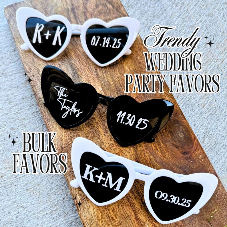 May include: Three pairs of heart-shaped sunglasses with white and black frames. The sunglasses have different text on each lens, including "R+K 07.14.25", "The Taylors 11.30.25", and "K+M 09.30.25".