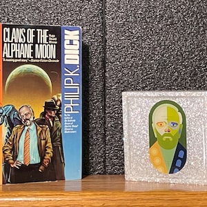 May include: A paperback book titled "Clans of the Alphane Moon" by Philip K. Dick. The book cover features a futuristic scene with a large, green insect and a man in a brown jacket. A small, square, white frame with a colorful portrait of a man inside is on a wooden surface.