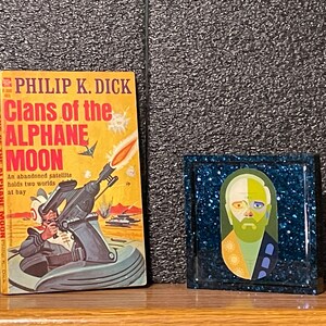 May include: A paperback book titled "Clans of the Alphane Moon" by Philip K. Dick with a yellow cover and a futuristic illustration of a spaceship.  The book is next to a small framed picture with a blue background and a colorful portrait of a person.