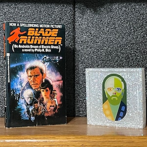 May include: A paperback copy of the science fiction novel "Blade Runner: Do Androids Dream of Electric Sheep?" by Philip K. Dick. The book cover features a futuristic cityscape with a man in a trench coat and a flying vehicle. The book is next to a small, white, square box with a colorful illustration of a person's face.