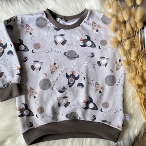 Könnte beinhalten: Ein hellgrauer Pullover mit braunem Saum. Der Pullover hat ein sich wiederholendes Muster aus Raketen, Planeten und dem Text "To the moon and back".