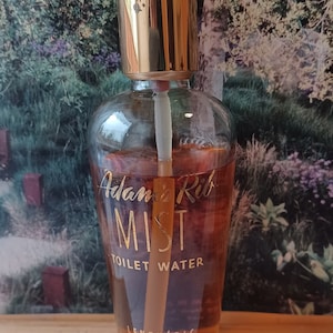 Puede incluir: Una botella de vidrio transparente con una tapa dorada y una etiqueta dorada que dice "Adam's Rib MIST TOILET WATER LENTHERIC NEW YORK PARIS LONDON CONT. 2 FL. OZ."