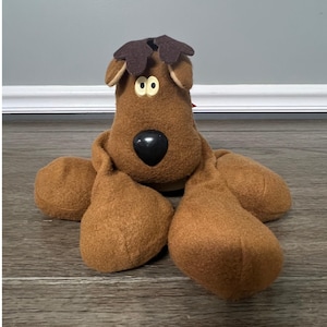 May include: A brown plush dog toy with a large black nose, yellow eyes, and brown felt antlers. The dog has large, floppy paws and is lying down. The toy is made of soft, textured fabric.