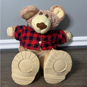 Op de afbeelding: Een bruine pluche teddybeer die een rood-zwart geruit overhemd en bruine laarzen draagt. De beer zit met gekruiste benen en heeft een bruine neus en bruine ogen. Op de voeten van de beer staat "The Original Care Bears" gedrukt.