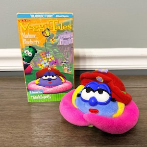 Puede incluir: Una cinta VHS de VeggieTales Madame Blueberry con una colorida portada animada y un juguete de peluche a juego con cuerpo azul, sombrero rojo y base rosa brillante, diseñado con grandes ojos blancos y labios rojos como en la película.