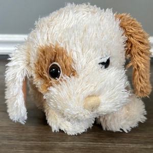 May include: A plush dog toy featuring shaggy white fur with light brown patches on the ears and around one eye. The stuffed animal has large, round, dark brown plastic eyes and a small, smooth, light tan fabric nose centered on its face.