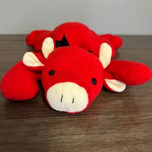 May include: A red plush toy shaped like a cow with a white muzzle and ears, and black eyes. The toy is lying on a wooden surface.