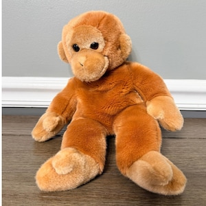 Puede incluir: Un mono de peluche naranja con una cara sonriente y ojos oscuros. El peluche tiene una textura suave y peluda y está sentado con los brazos y las piernas extendidos. Las manos y los pies del mono son de un tono naranja más claro.