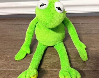 Könnte beinhalten: Ein leuchtend hellgrünes Plüschfrosch-Spielzeug mit weicher, flauschiger Textur. Der Frosch hat große, runde weiße Augen mit schwarzen Pupillen, lange, schlanke Arme und lange Beine mit Schwimmfüßen. Die Figur hat eine einfache, abgerundete Kopf- und Körperform.