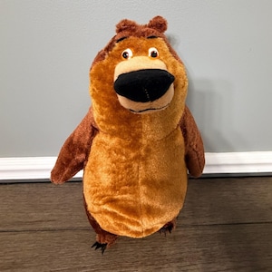 May include: A soft toy bear in shades of brown and tan. The bear features a large black nose, brown arms, and small black claws. It is standing upright and has a friendly expression.