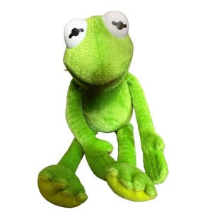 Könnte beinhalten: Ein leuchtend grünes Frosch-Stofftier mit weicher, flauschiger Textur. Die Figur hat große, weiße, ovale Augen mit schwarzen Pupillen, einen breiten Kopf, lange, schlanke Arme und lange Beine mit abgerundeten Füßen. Das Material ist lindgrün.