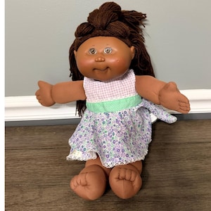 May include: A doll with dark brown yarn hair styled in a top knot, featuring a round face, brown skin, and hazel eyes. The doll wears a dress with a light purple and white gingham bodice, a mint green waistband, and a purple floral skirt.