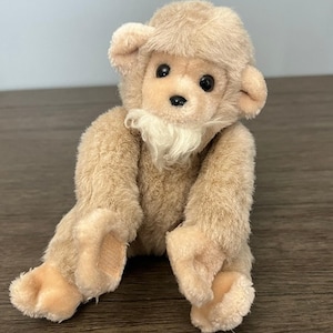 May include: A plush monkey toy with a light brown fur body, a matching head covering, and a white beard. The monkey has black eyes and tan hands and feet. The toy is sitting on a wooden surface.