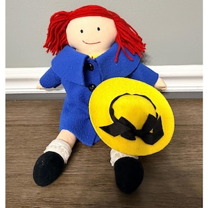 May include: A fabric doll with bright red yarn hair, wearing a blue coat with two black buttons over a yellow shirt. The doll has black shoes with white lace-trimmed socks and is holding a yellow felt hat featuring a black ribbon bow.