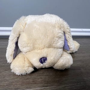 May include: A plush, cream-colored dog toy with floppy ears and a purple nose and ear accents. The soft toy is lying down on a wooden surface, with its head resting on its paws. The toy is designed to resemble a dog.