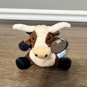 May include: A plush toy of a longhorn steer. The toy has a white face and horns, brown and white body, and black hooves. It has a tag attached to its ear.