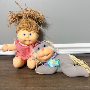 Puede incluir: Una muñeca Cabbage Patch Kids con cabello de lana marrón y ojos azules que viste un vestido de cuadros rojos y blancos, junto a un juguete de peluche de foca gris con cabello de lana rubio, un collar morado y una etiqueta azul con forma de pez.