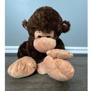 May include: A plush monkey toy with brown fur and a tan face, hands, and feet. The stuffed animal has large, floppy feet and is sitting down. The monkey's fur is soft and fluffy, and it has a friendly expression.