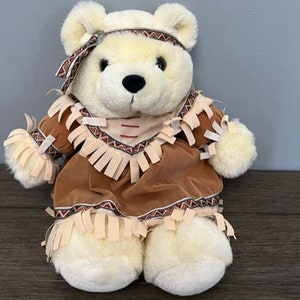 May include: A white plush teddy bear wearing a brown and tan Native American costume with fringe and beading.