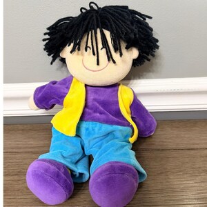 May include: A plush doll with a cream-colored face, black yarn hair, and a smiling expression. The doll wears a purple shirt, a yellow vest, and blue pants. The doll's feet are purple.