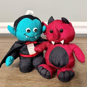 May include: Two plush Halloween figures: a teal vampire with a black cape and a red devil with black accents. The vampire has a smiling face with fangs. The devil has horns and a black belly. Both are sitting on a wooden surface.