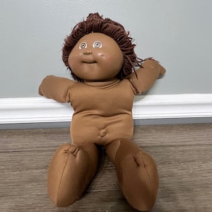 May include: A vintage Cabbage Patch Kid doll with brown skin, a round face, and brown yarn hair. The doll is wearing a brown fabric body and has open arms and legs. The doll is sitting on a wooden floor.